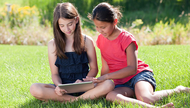 Two girls reading on a tablet in the grass.