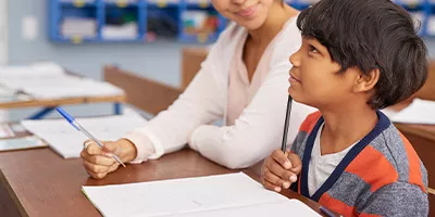 a teacher supporting a student sitting at a desk in a classroom 