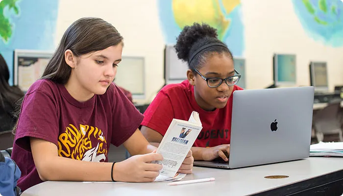 Students in a classroom reading a book and working on a laptop, engaged in learning and independent reading activities.