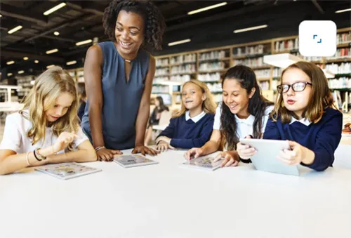 Teacher standing over a group of students seated at a table who are focused on reading books together in a bright, inviting library.
