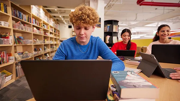Student sitting at a library table, focused on a laptop, with two other students working on laptops in the background