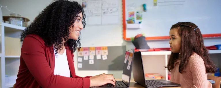 A teacher on a laptop with a young student in a classroom 